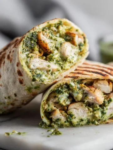 Overhead homemade pesto chicken wrap recipe cut in half on a white marble counter with crumbs and pesto smudges, casual kitchen look.