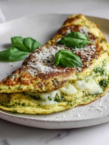 Overhead shot of a homemade pesto omelette recipe folded on a white plate with basil and parmesan, on a white kitchen counter.