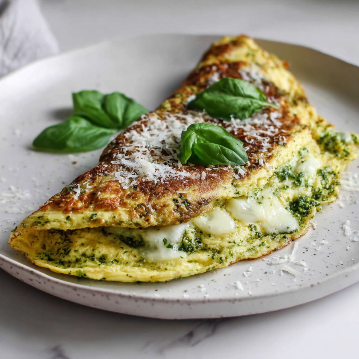 Overhead shot of a homemade pesto omelette recipe folded on a white plate with basil and parmesan, on a white kitchen counter.