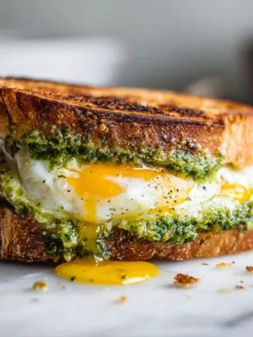 Homemade pesto breakfast sandwich recipe with eggs and mozzarella on sourdough, overhead shot on white marble kitchen counter.