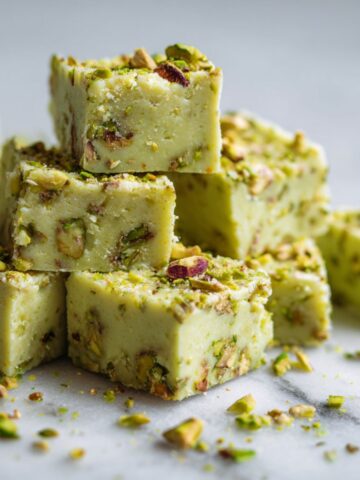 Overhead photo of homemade pistachio fudge recipe squares on a white marble kitchen counter, cut into rustic pieces with pistachios sprinkled on top.