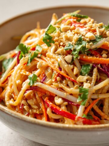 Overhead view of a bowl of homemade spicy Thai peanut noodles recipe topped with vegetables, crushed peanuts, and fresh herbs on a white kitchen counter.