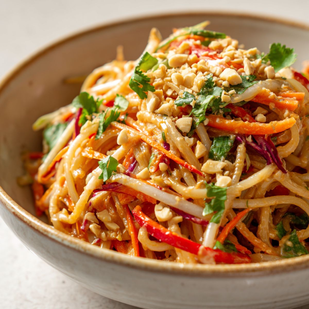 Overhead view of a bowl of homemade spicy Thai peanut noodles recipe topped with vegetables, crushed peanuts, and fresh herbs on a white kitchen counter.