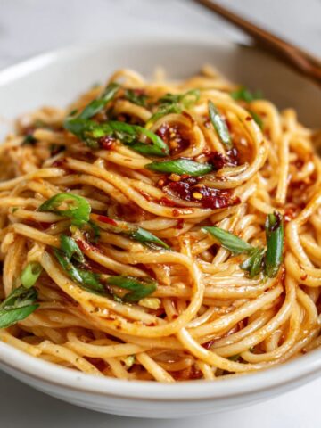 Overhead photo of spicy Szechuan noodles recipe in a bowl on a white kitchen counter, garnished with chili oil and green onions, looking homemade and rustic.