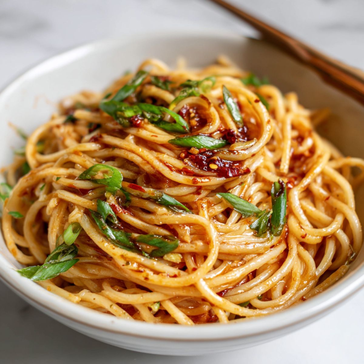 Overhead photo of spicy Szechuan noodles recipe in a bowl on a white kitchen counter, garnished with chili oil and green onions, looking homemade and rustic.