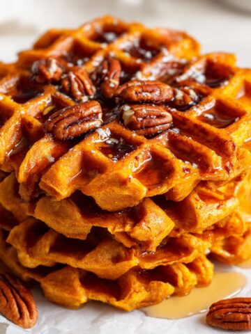 Homemade sweet potato waffles recipe stacked on a white kitchen counter with maple syrup and pecans, photographed from above.