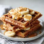 Homemade peanut butter banana waffles recipe stacked on a plate, drizzled with peanut butter and topped with sliced bananas on a white kitchen counter.