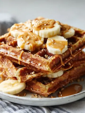 Homemade peanut butter banana waffles recipe stacked on a plate, drizzled with peanut butter and topped with sliced bananas on a white kitchen counter.