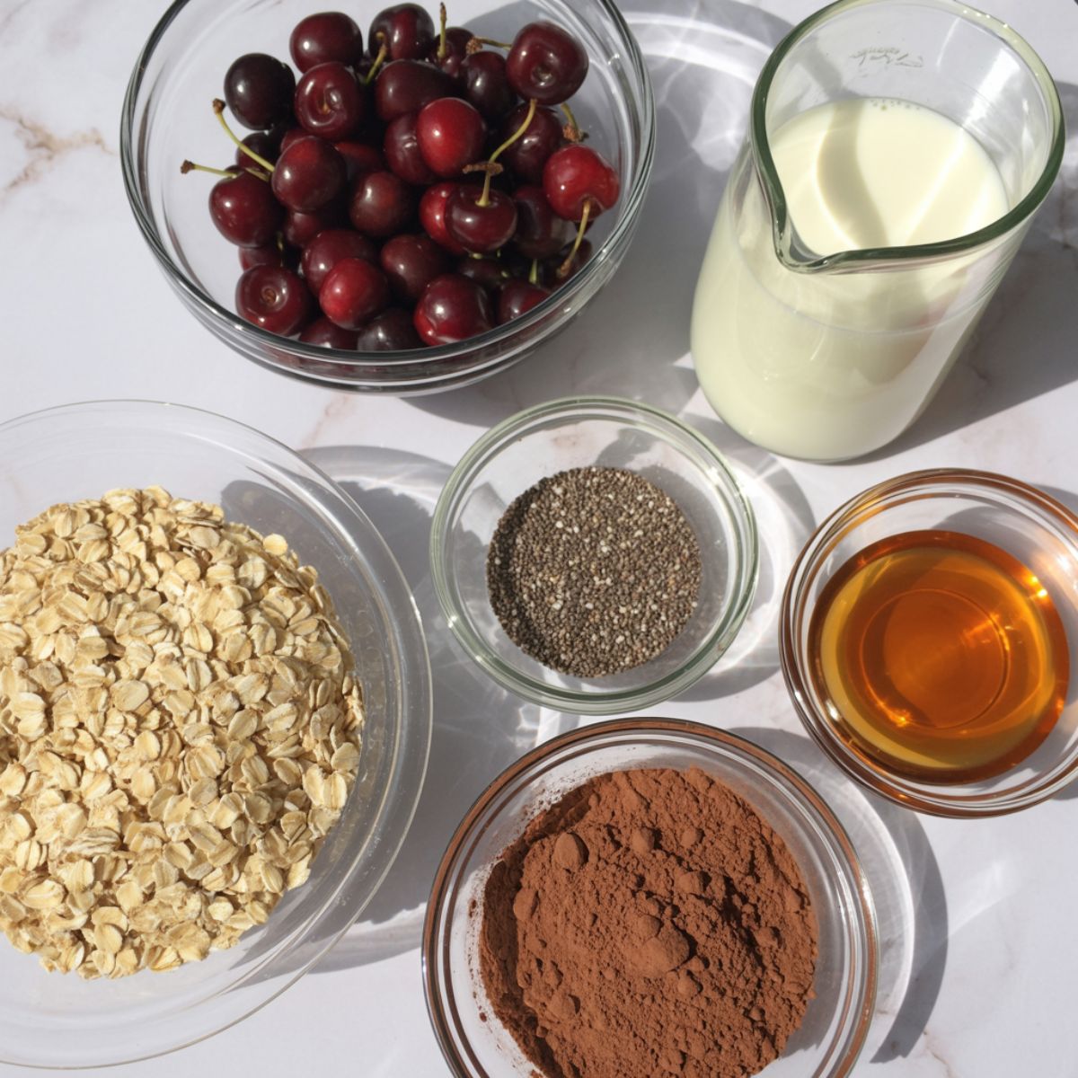Overhead shot of rolled oats, cherries, cocoa powder, chia seeds, almond milk, and maple syrup arranged casually on a white kitchen counter.