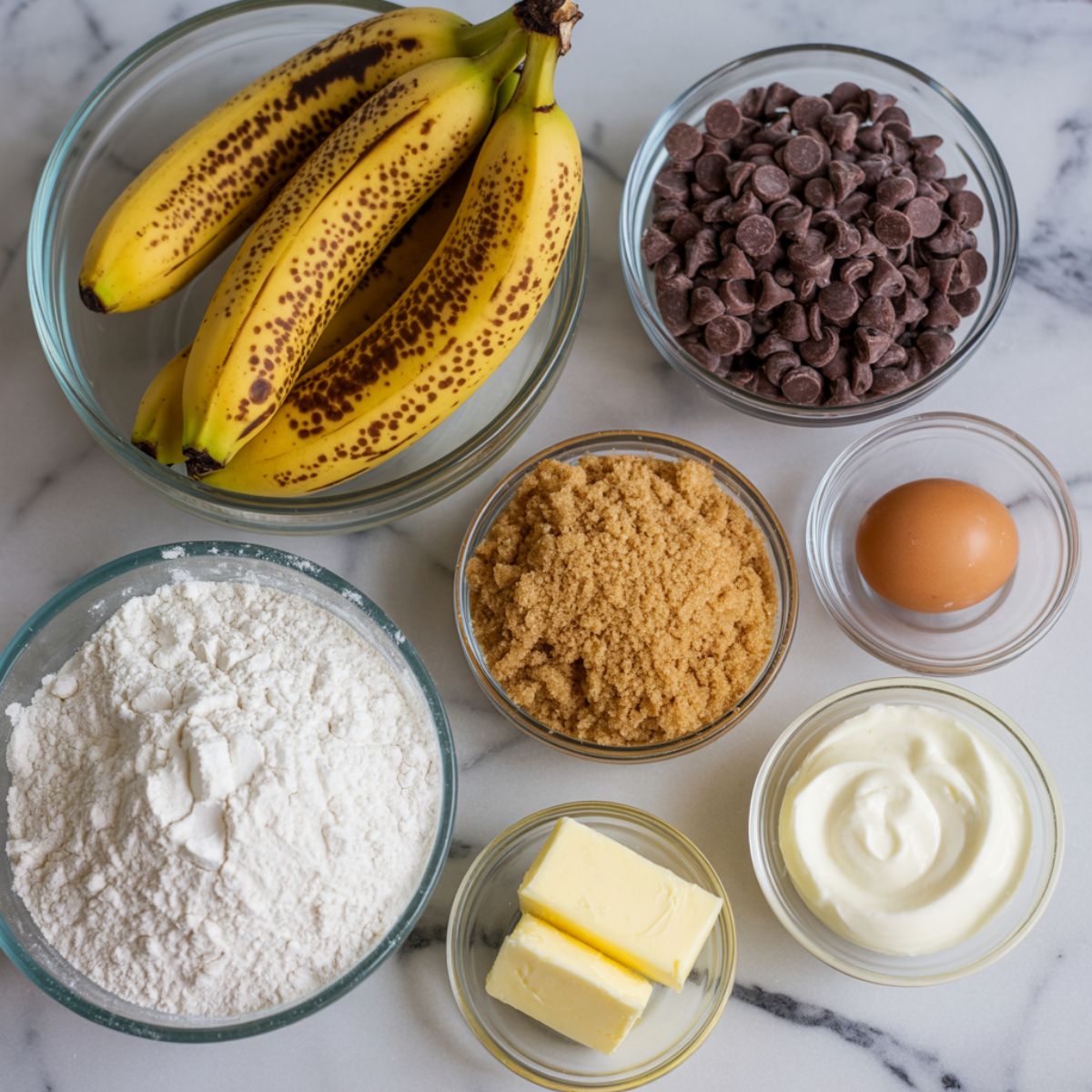 Overhead view of ripe bananas, chocolate chips, flour, brown sugar, egg, butter, and sour cream on a white kitchen counter for banana bread ingredients.