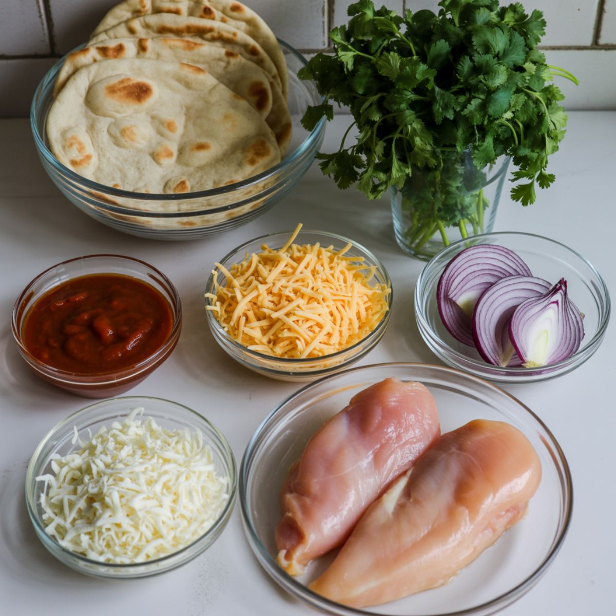 Flatlay of BBQ chicken flatbread pizza ingredients including naan, barbecue sauce, cheese, chicken breast, red onions, and cilantro on a white kitchen counter.
