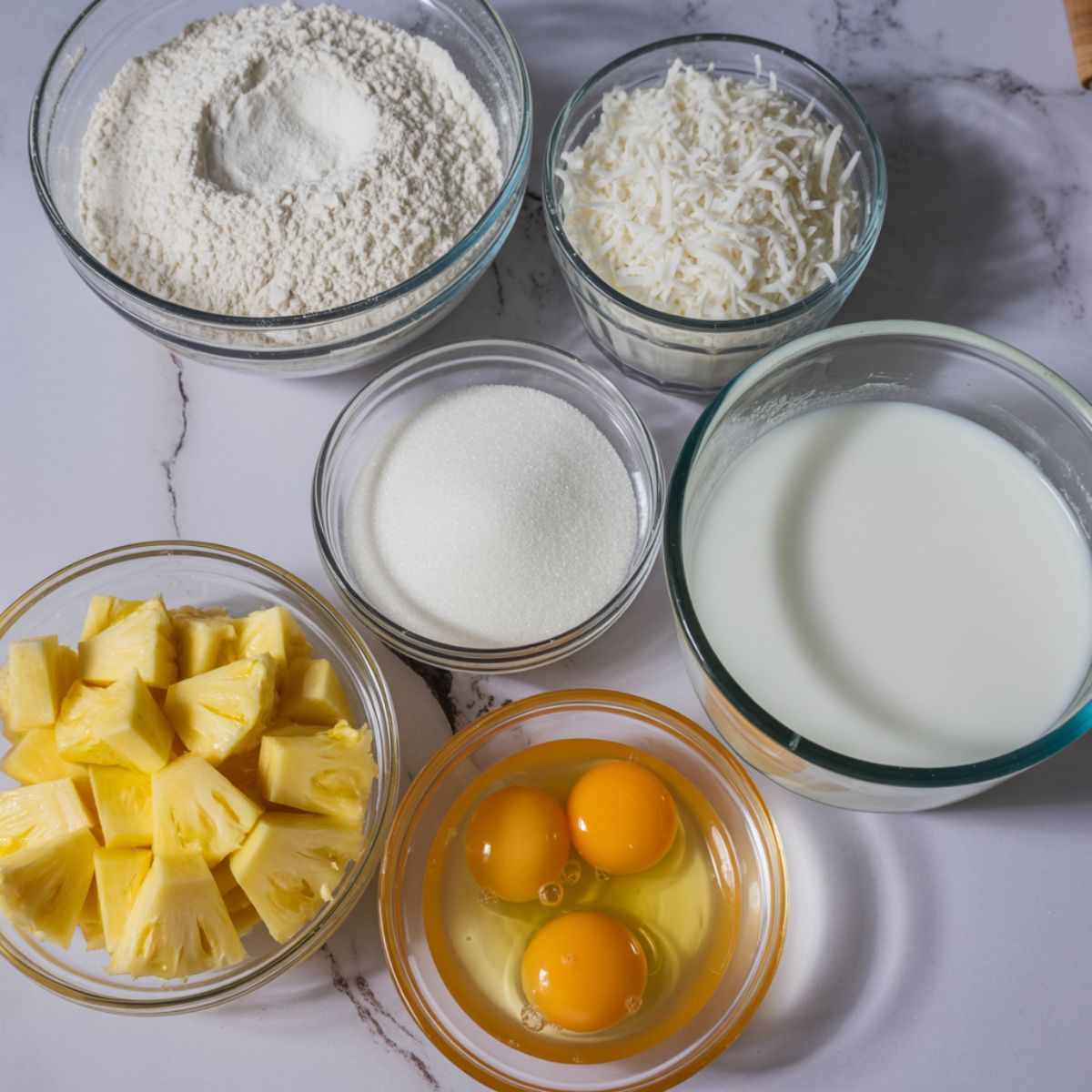 Overhead photo of coconut pineapple cupcake ingredients including flour, shredded coconut, pineapple chunks, eggs, and coconut milk on a white kitchen counter.