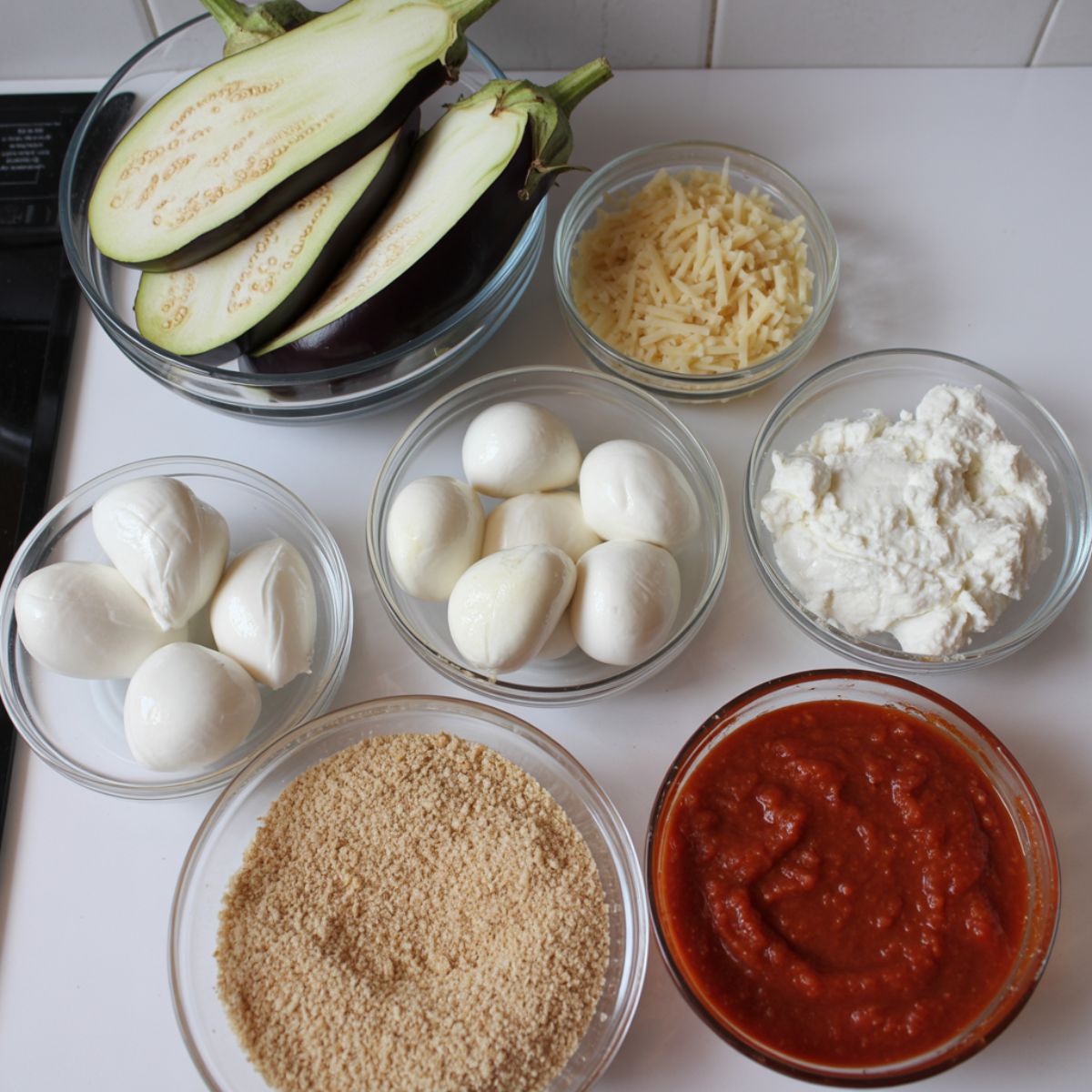 Overhead view of eggplant parmesan casserole ingredients including eggplant slices, mozzarella, ricotta, parmesan, breadcrumbs, and marinara sauce on a white kitchen counter.