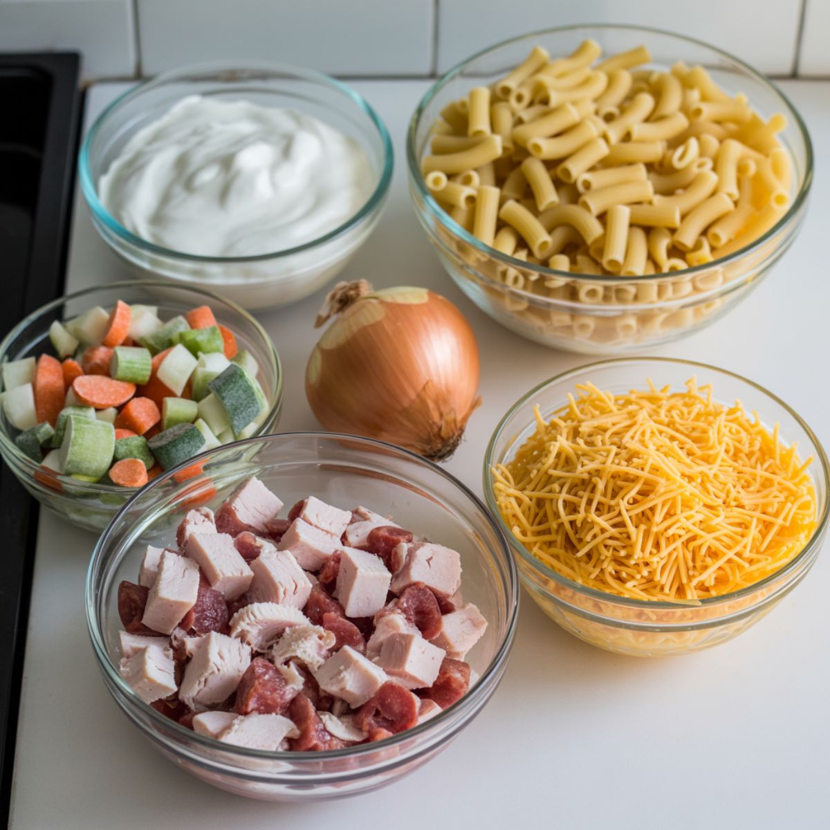 Flat lay of ingredients for cheesy turkey casserole including diced turkey, pasta, shredded cheese, sour cream, canned soup, and vegetables on a white kitchen counter.