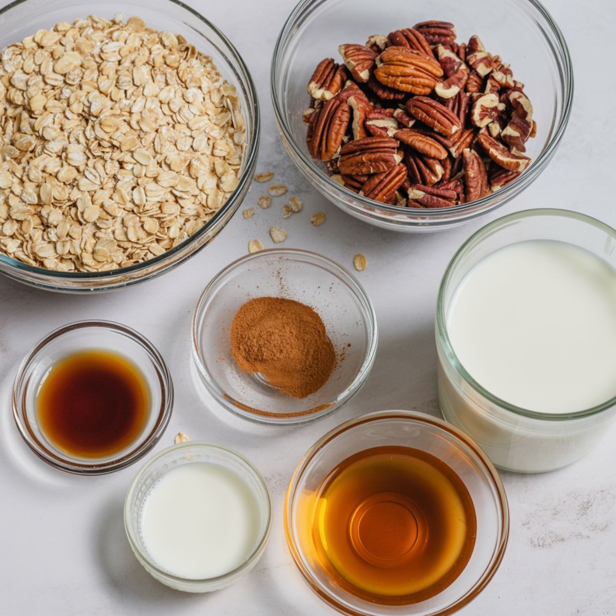 Overhead view of pecan pie overnight oats ingredients including oats, pecans, cinnamon, vanilla, maple syrup, and milk on a white counter.