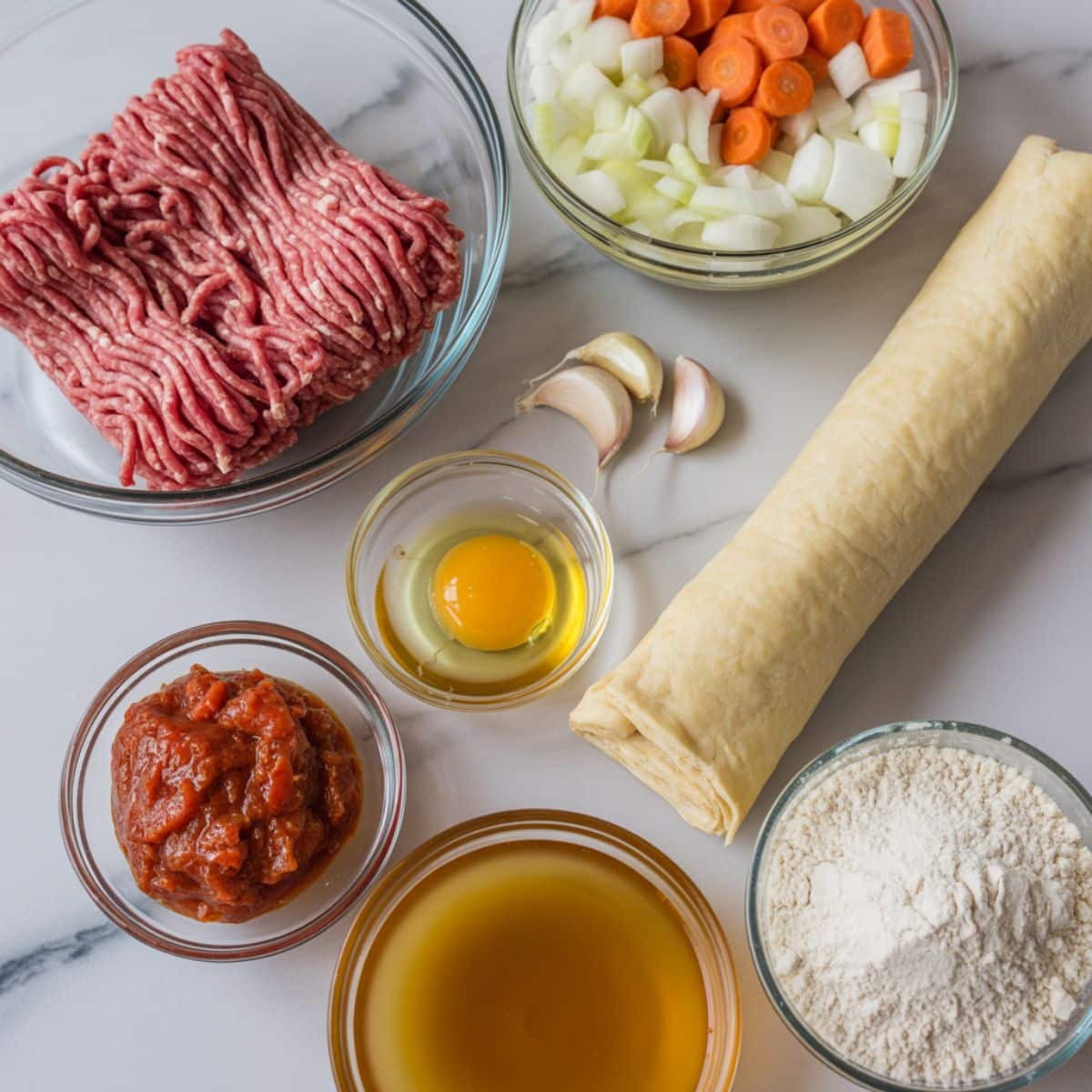 Overhead shot of minced beef pie ingredients including ground beef, onions, carrots, garlic, puff pastry, and stock on a white marble counter.