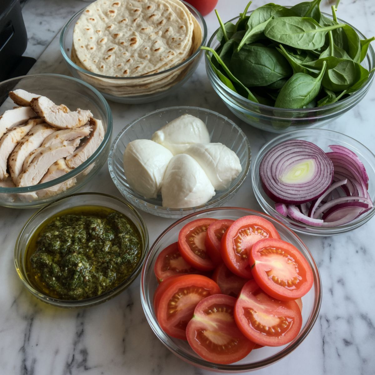 Overhead shot of pesto chicken wrap ingredients—tortillas, chicken strips, mozzarella, pesto, spinach, tomatoes, and red onion on a white kitchen counter in a casual homemade style.