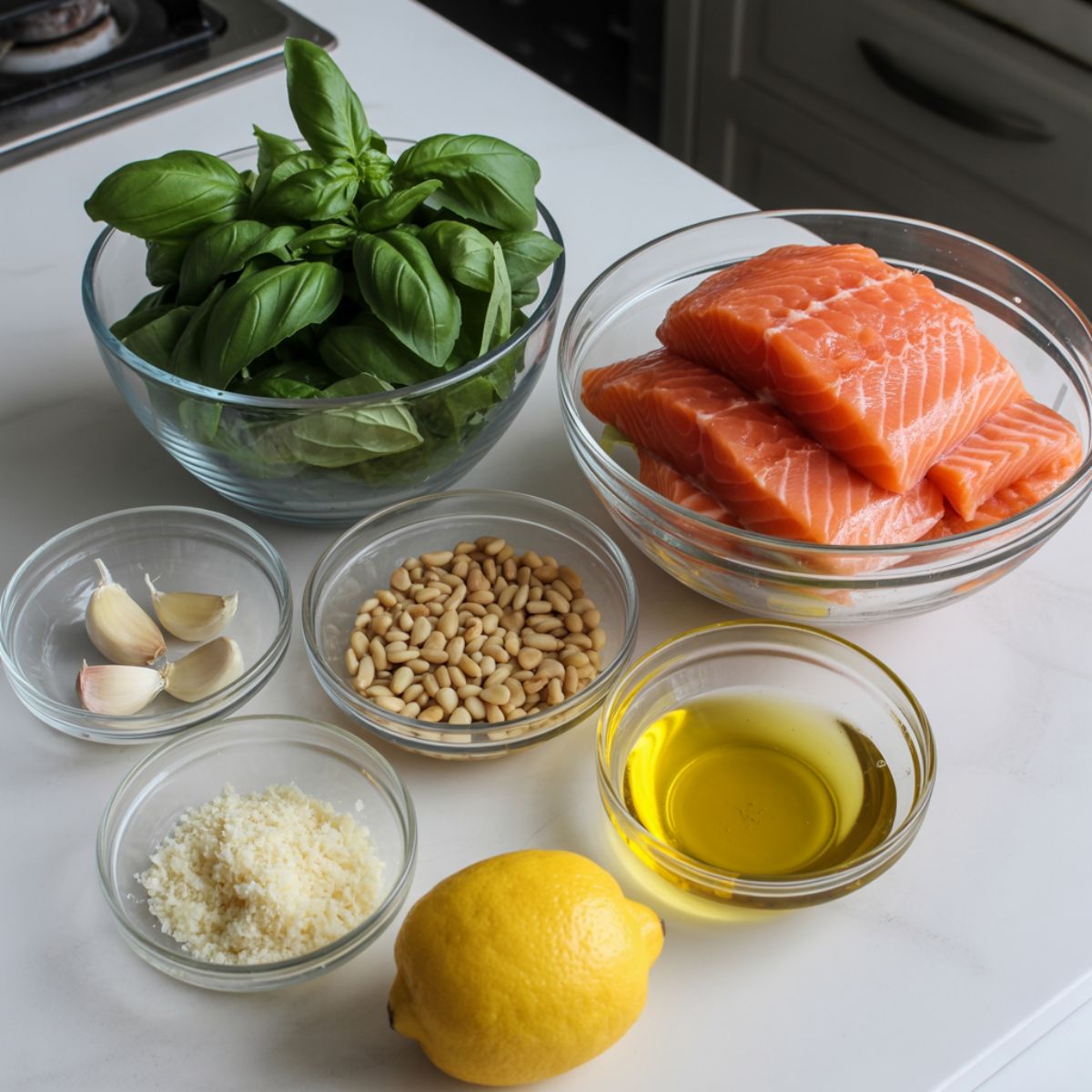 Raw ingredients for pesto salmon recipe including salmon fillets, fresh basil, garlic, pine nuts, parmesan, olive oil, and lemon on a white kitchen counter.