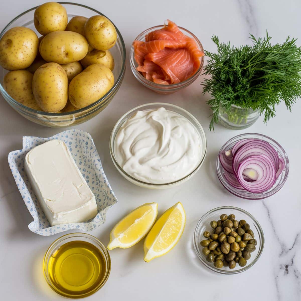 Flat lay of ingredients for smoked salmon potato recipe including potatoes, smoked salmon, dill, red onion, sour cream, cream cheese, lemon, and capers on a white marble counter.”