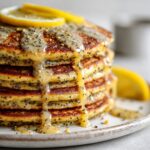 Stack of fluffy lemon poppy seed pancakes recipe drizzled with lemon glaze on a white kitchen counter, photographed from above in a homemade style.