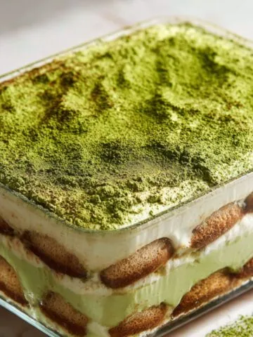 Homemade matcha tiramisu recipe in a glass dish with green matcha layers and light dusting of matcha powder on a white kitchen counter, photographed from above.