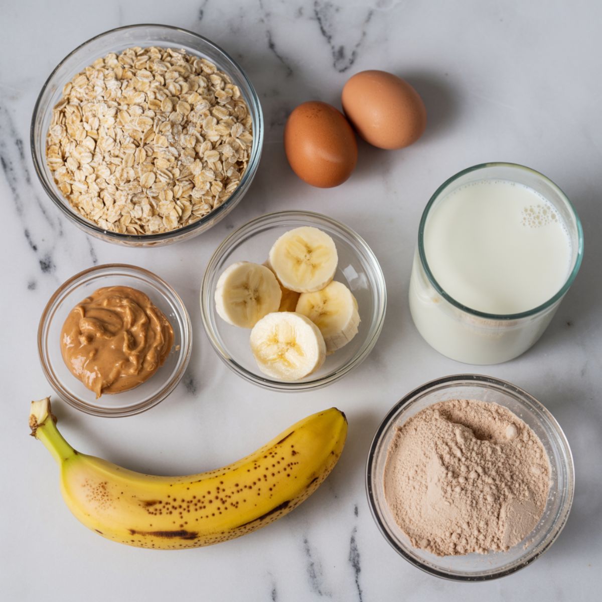 Overhead view of peanut butter protein pancakes ingredients including oats, eggs, peanut butter, banana, protein powder, and almond milk on a white kitchen counter in a homemade casual style.