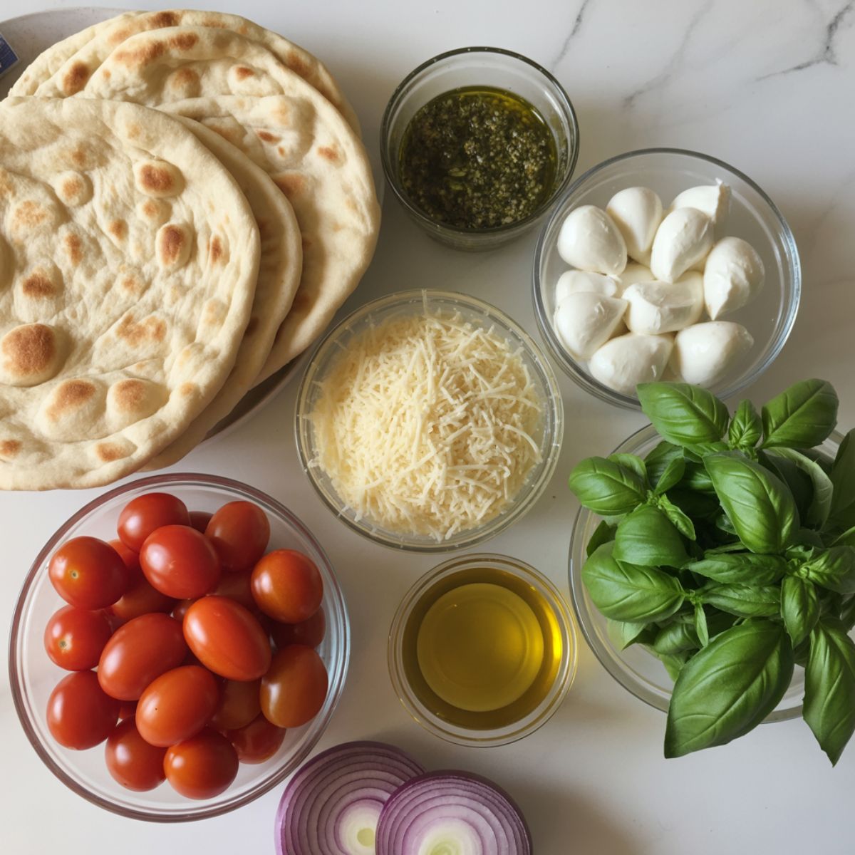 Flatbread pizza ingredients including pesto, mozzarella, parmesan, cherry tomatoes, onion, and basil on a white kitchen counter, photographed from above.