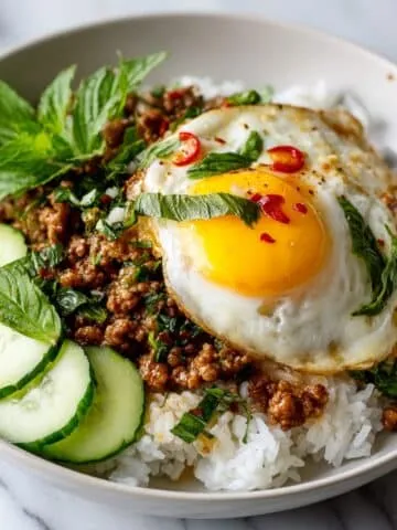 Homemade Thai basil minced pork recipe served over jasmine rice with a fried egg on top, garnished with basil and cucumber slices, photographed from above on a white kitchen counter.