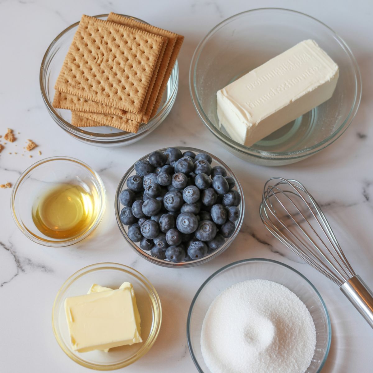 Overhead shot of real cheesecake ingredients — graham crackers, blueberries, cream cheese, and butter on a white kitchen counter, slightly messy homemade setup.