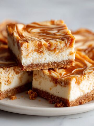 Overhead view of homemade dulce de leche cheesecake bars recipe on a white marble counter with gooey caramel swirls and a few crumbs around.