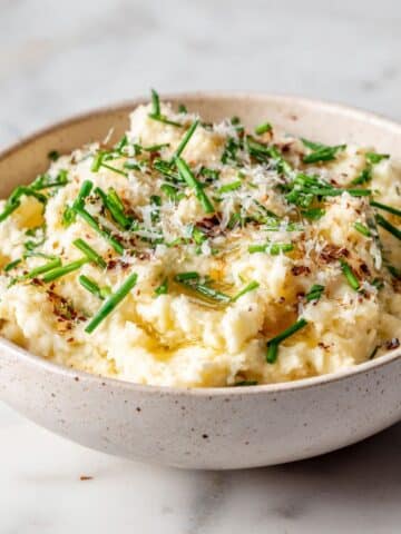 Homemade truffle mashed potatoes recipe in a bowl, topped with chives and Parmesan, photographed on a white marble counter from above.