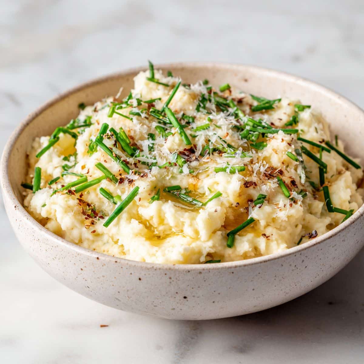 Homemade truffle mashed potatoes recipe in a bowl, topped with chives and Parmesan, photographed on a white marble counter from above.