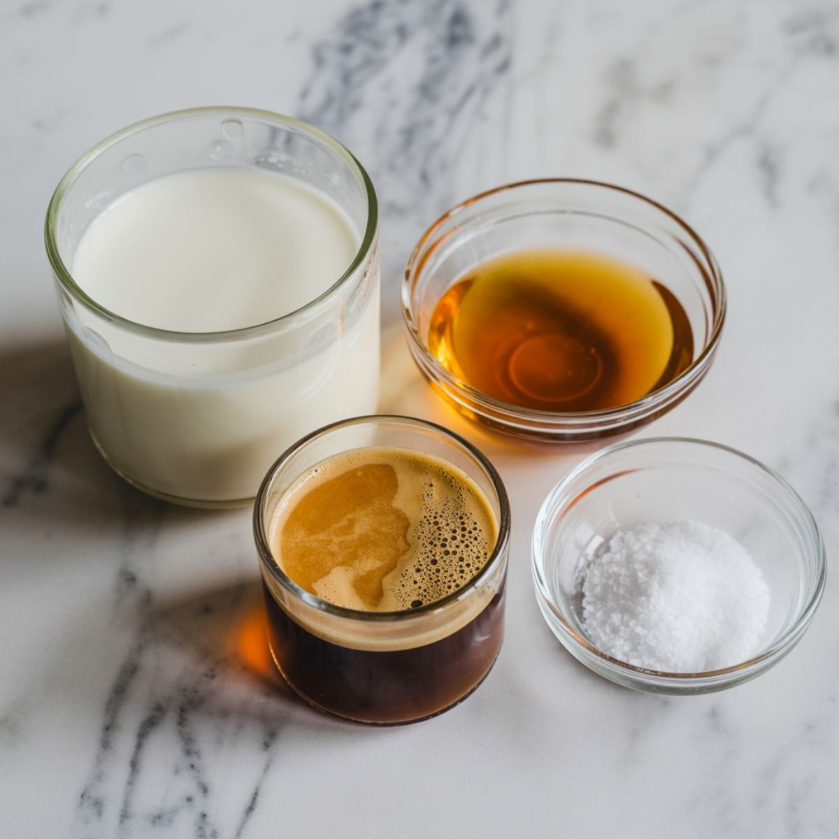 Overhead shot of raw ingredients for honey almond milk flat white including almond milk, honey jar with dipper, espresso shot, and pinch bowl of sea salt on a white marble kitchen counter.