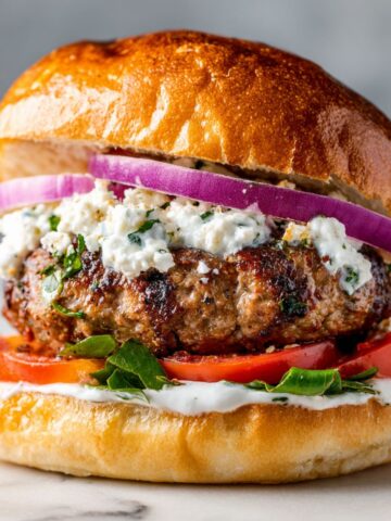 Homemade Mediterranean lamb burger recipe with feta, tzatziki, and fresh herbs on a white marble counter, captured from above in natural light.