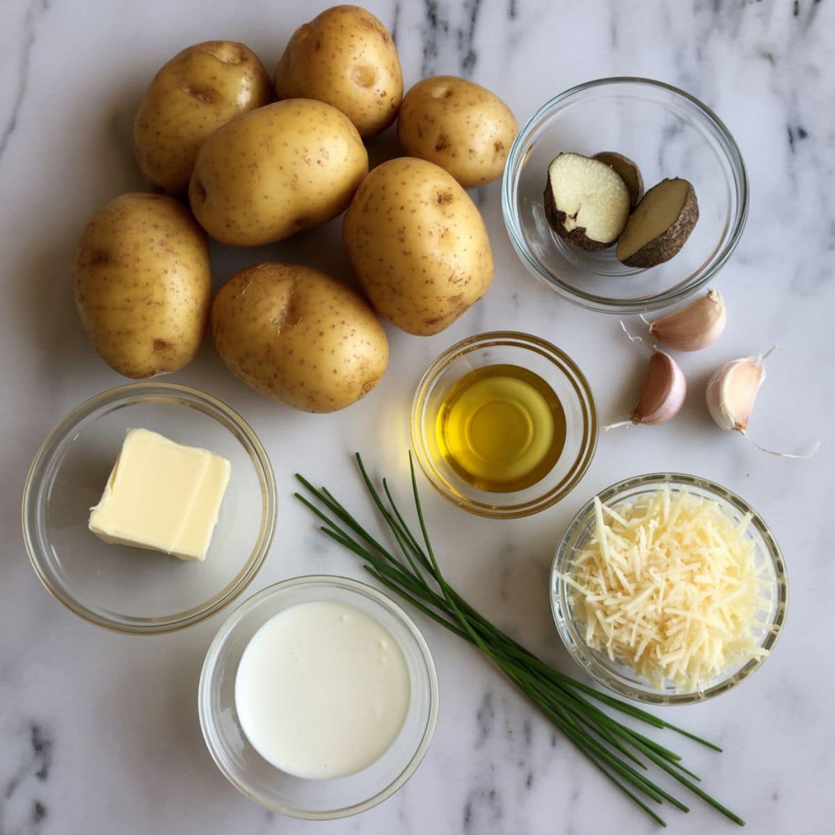 Overhead photo of truffle mashed potato ingredients on a white marble counter including potatoes, truffle oil, butter, cream, garlic, and chives.