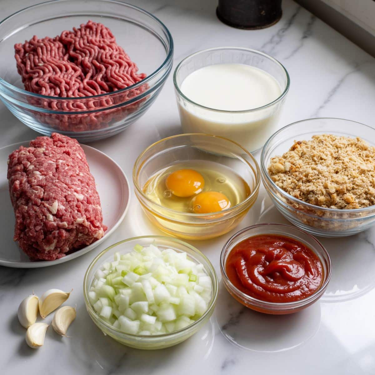 Overhead view of meatloaf cups ingredients including ground beef, breadcrumbs, eggs, milk, onion, garlic, and ketchup glaze on a white kitchen counter.