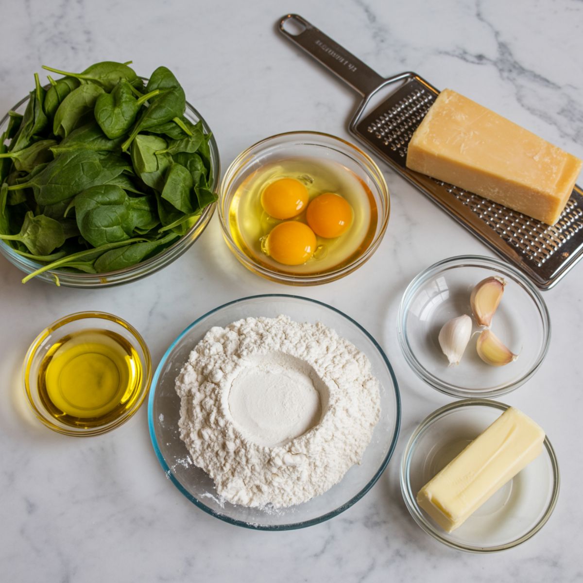 Top-down view of fresh spinach, eggs, flour, garlic, butter, and parmesan laid out on a marble counter for homemade tagliatelle.