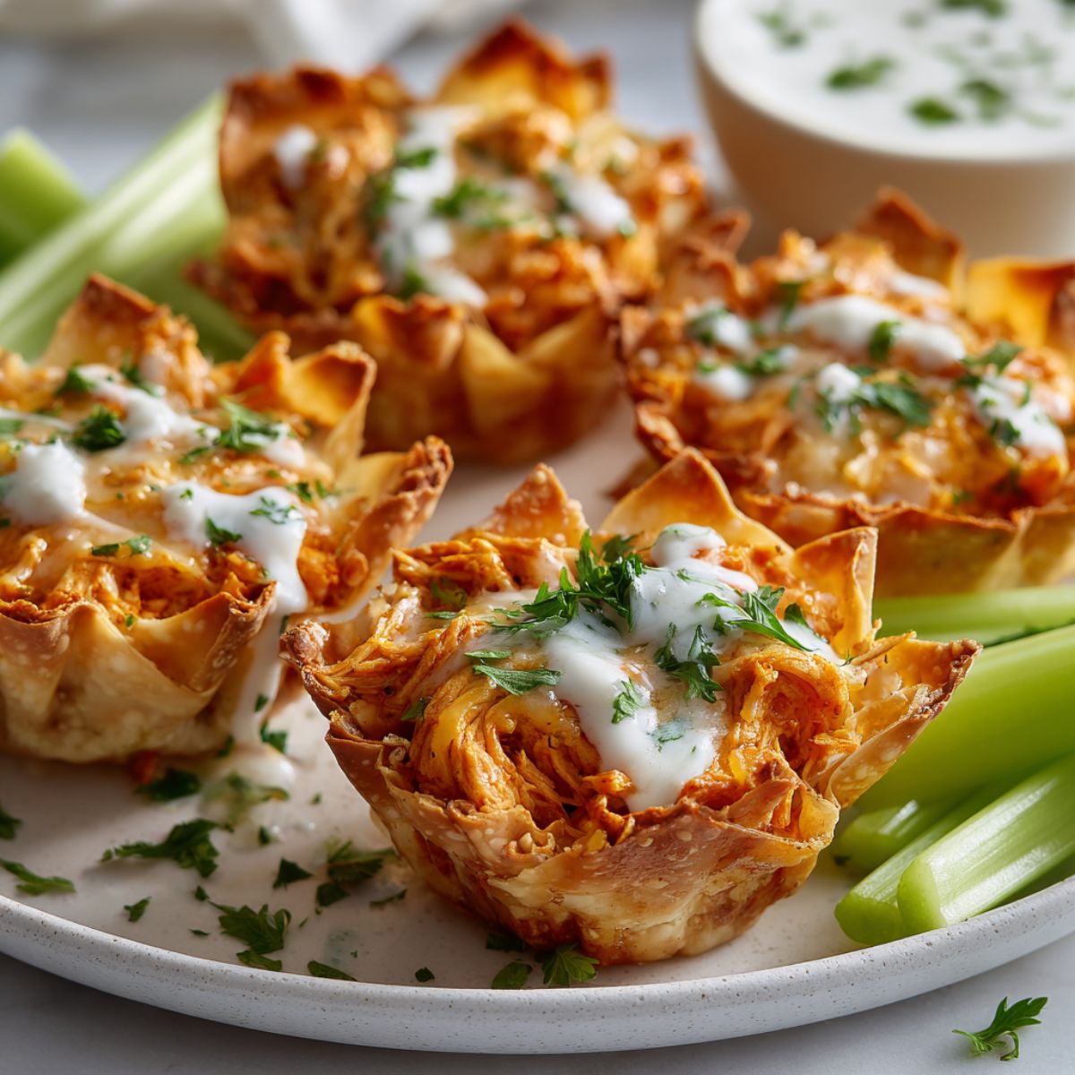 Homemade buffalo cups recipe on a white kitchen counter, topped with melted cheese, ranch drizzle, and parsley — a cozy game day appetizer served with celery sticks.