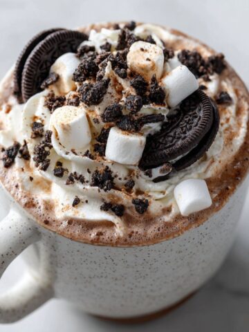 Homemade cookies and cream hot cocoa recipe topped with whipped cream, crushed Oreos, and marshmallows on a white kitchen counter – overhead shot with cozy homemade vibe.
