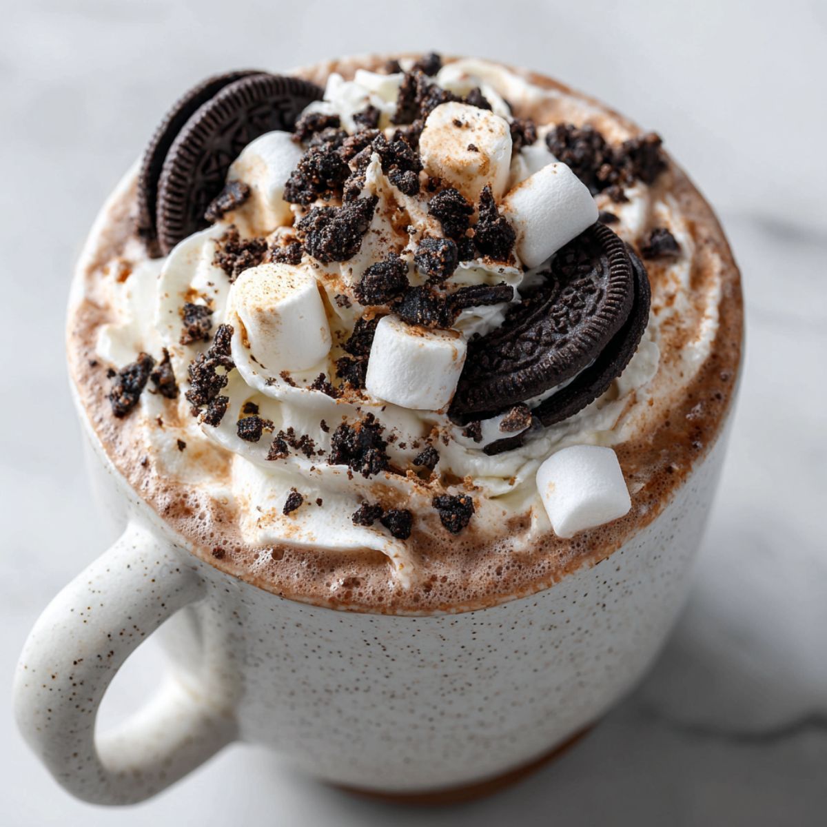 Homemade cookies and cream hot cocoa recipe topped with whipped cream, crushed Oreos, and marshmallows on a white kitchen counter – overhead shot with cozy homemade vibe.