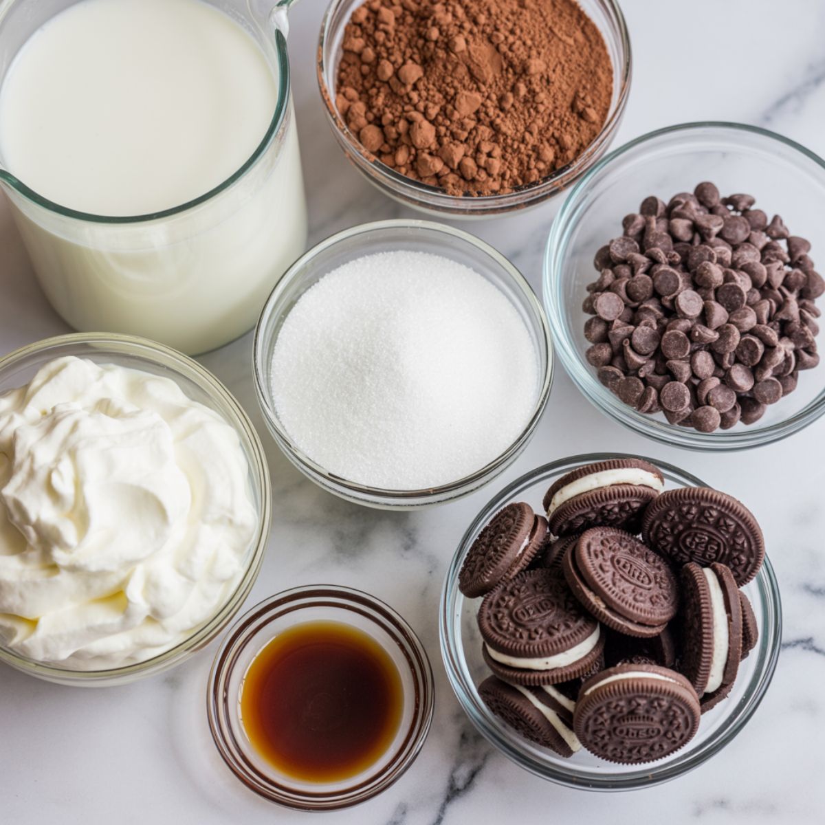 Flat lay of ingredients for cookies and cream hot cocoa – milk, cocoa powder, sugar, chocolate chips, vanilla extract, and crushed Oreos on a white marble kitchen counter.