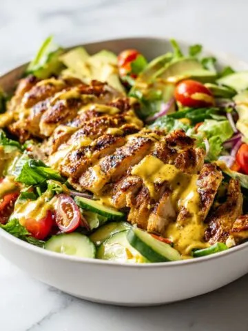 Homemade honey mustard chicken salad recipe in a white bowl with grilled chicken, greens, avocado, and honey mustard dressing on a white kitchen counter.