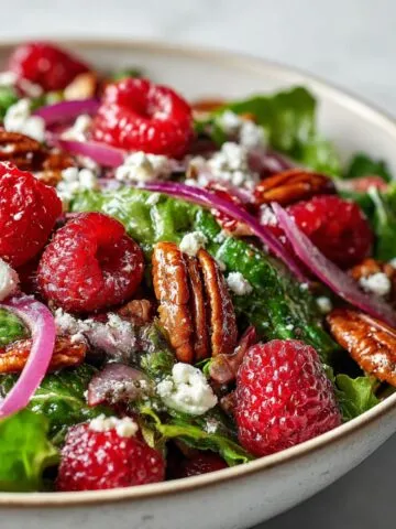Homemade raspberry salad recipe with fresh greens, raspberries, feta, and pecans on a white kitchen counter, photographed overhead.