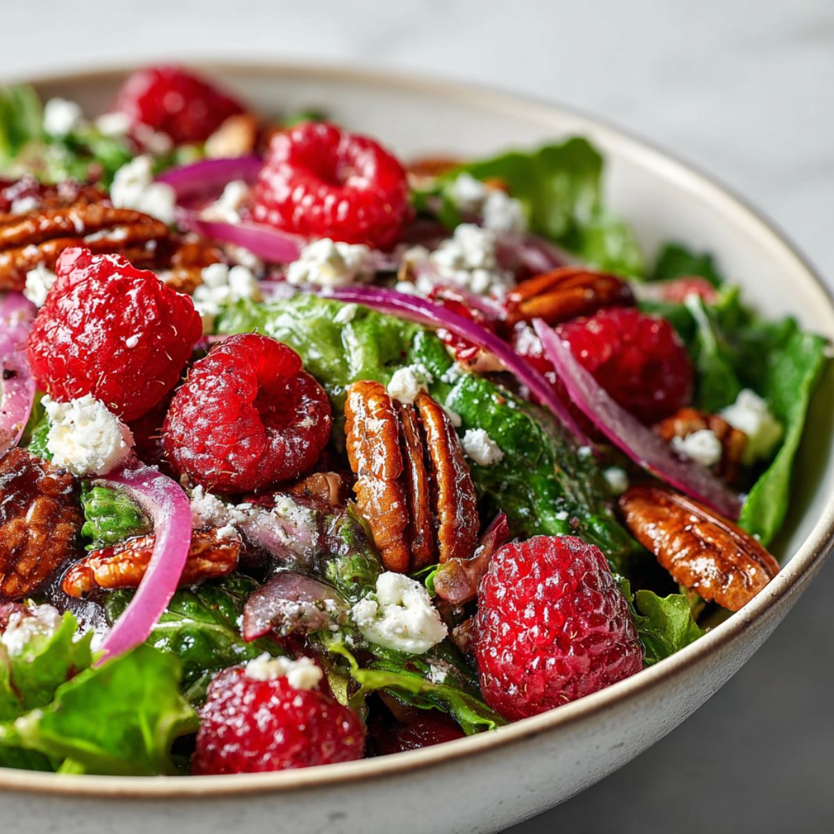 Homemade raspberry salad recipe with fresh greens, raspberries, feta, and pecans on a white kitchen counter, photographed overhead.