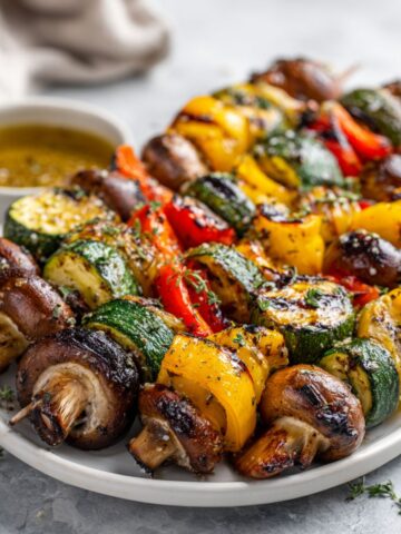 Overhead shot of homemade grilled vegetable skewers recipe on a white kitchen counter with herbs and marinade bowl.