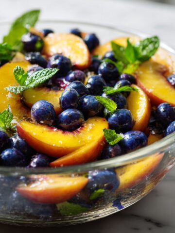 Overhead photo of a homemade blueberry peach salad recipe with sliced peaches, fresh blueberries, and mint leaves in a glass bowl on a white marble kitchen counter.