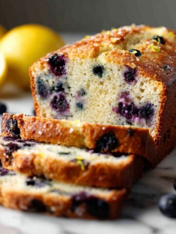 Homemade blueberry lemon bread recipe loaf sliced on a white marble counter with fresh blueberries and lemon zest visible.