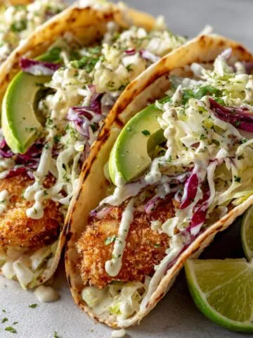 Overhead shot of homemade crispy fish tacos recipe on a white kitchen counter with cabbage slaw, avocado, and lime, showing a natural, homemade feel.
