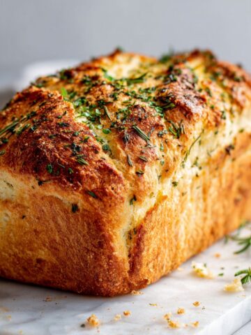 Homemade garlic herb bread recipe loaf with golden crust and fresh herbs on a white marble kitchen counter.