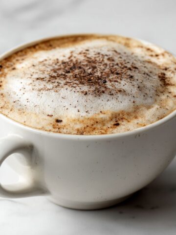Overhead view of a homemade hazelnut cappuccino recipe with frothy foam and a light cinnamon sprinkle on a white marble kitchen counter.